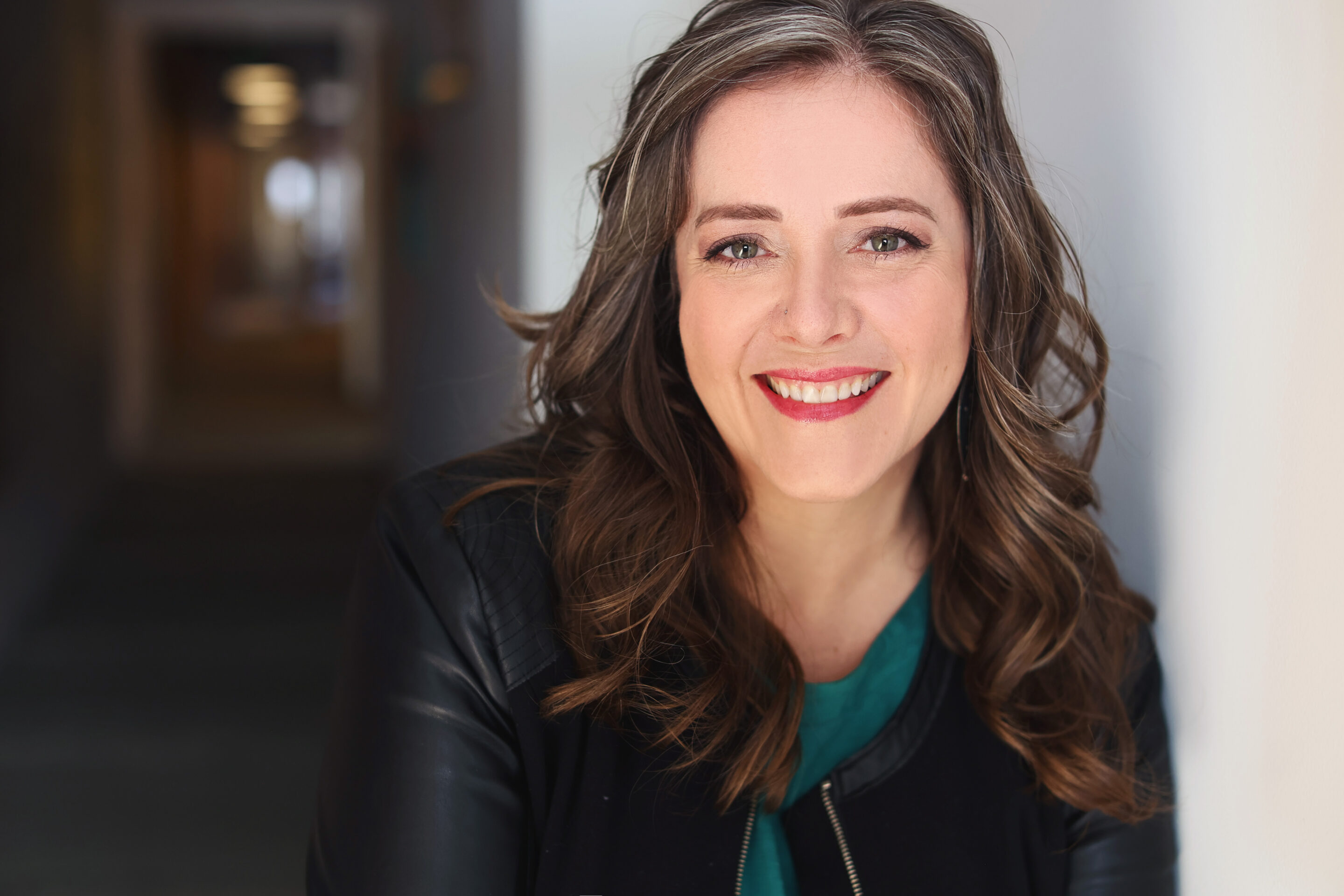 Angella Johnson in a green shirt and black jacket leaning against a white wall with a hallway and business offices behind her.