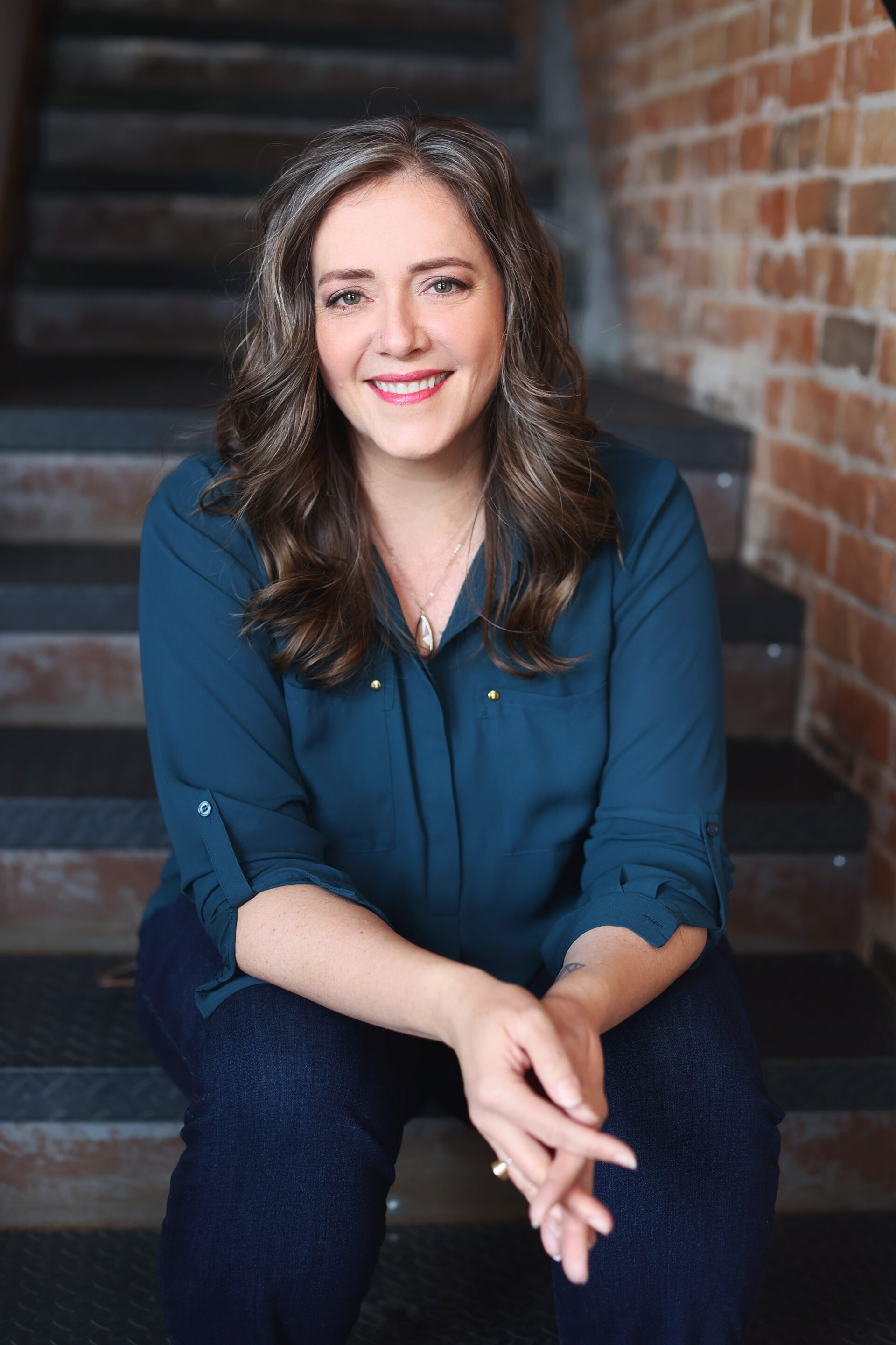 Angella sitting on stairs wearing a dark teal green shirt with her hands folded slightly in front of her.